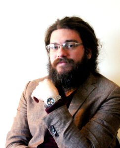 A bearded man with glasses and long hair wearing a brown jacket